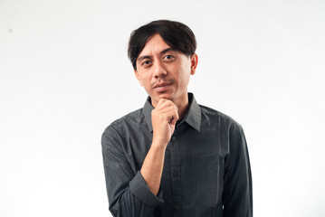 An Asian man in a gray shirt, standing with his hand on his chin, looking thoughtful or pondering....
