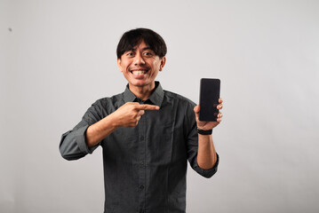 An Asian man in a gray shirt smiling broadly while pointing to a smartphone he holds in his other...