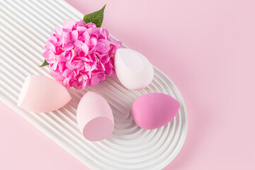 Beautiful beauty blender sponges for applying liquid cosmetics on a white embossed podium with flower. top view. Makeup tool. pink background. closeup
