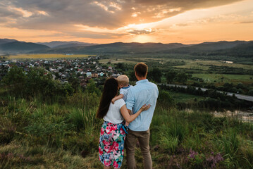 Mom, dad embrace baby looking at sunset. Happy family holiday on summer day. Mother, father, baby  in mountains. Parents hugging child. Spend time together. Back view.