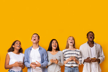 Group of surprised multinational students with smartphones looking at empty space over yellow...