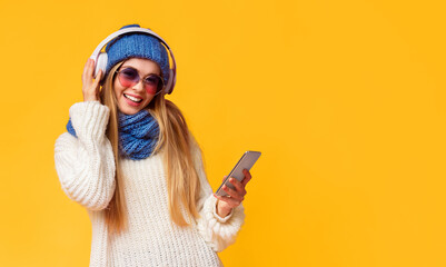 New music app for mobile phone. Blonde winter girl in eyewear listening to music, using smartphone and wireless headset, pink studio background