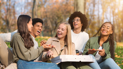 Joyful multiracial teen friends laughing and eating pizza, having picnic in forest