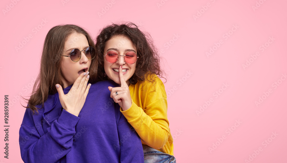 Wall mural Two friends wearing colorful outfits, one whispering a secret while the other looks surprised. Fun and playful moment captured on a pink background.