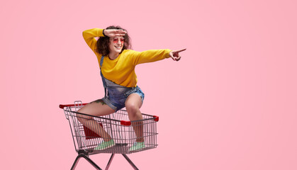 Cheerful woman in casual outfit, sitting in a shopping cart and pointing forward. Fun and playful...