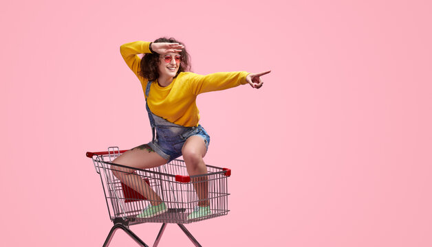 Cheerful woman in casual outfit, sitting in a shopping cart and pointing forward. Fun and playful concept against a bright pink background.