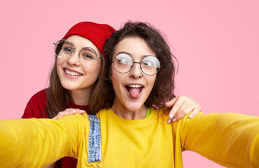 Two young friends wearing glasses taking a fun selfie together, one sticking out her tongue, against a bright pink background.