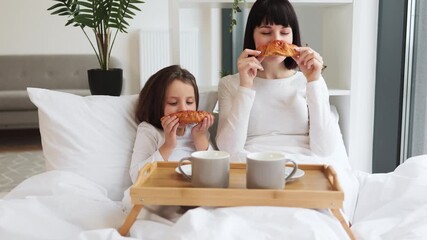 Mother and daughter eating croissants together in bed, sharing quality family time. Wooden breakfast tray with coffee mugs visible. Bright and cozy bedroom. - Powered by Adobe