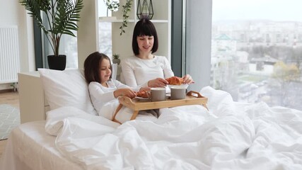 Mother and daughter sharing breakfast in bed, smiling at each other while holding croissants, bonding over a cozy morning. Wooden tray with croissants and cups on bed creates warm, intimate atmosphere - Powered by Adobe