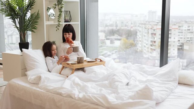 Mother and daughter enjoying breakfast in bed with croissants and coffee. Cozy morning by window with city view and indoor plants. Bonding time for family filled with warmth and happiness.