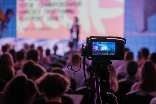 Video camera records live conference attended by audience in auditorium, capturing event for streaming or archival purposes.