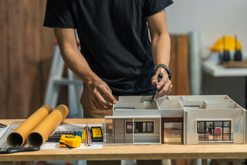 A modern engineer's desk featuring a detailed house model, blueprints, and renovation plans. The wooden desk is neatly organized in a bright, glass-walled room, with no people present.