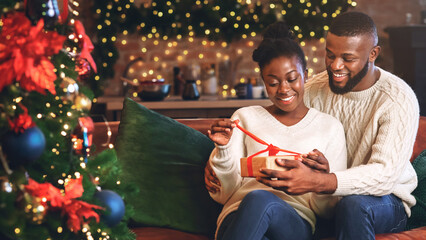 Happy african american woman unwrapping her christmas present with husband, celebrating holidays together at home.