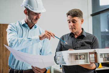 A Caucasian middle-aged male engineer contractor and an Italian architect engineer are seated at a desk, discussing renovation plans with blueprints and a house model in front of them.