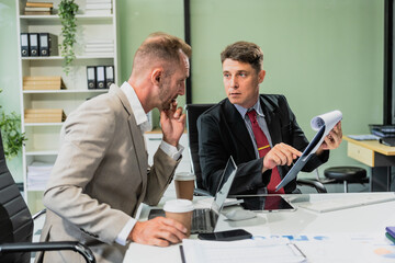 Caucasian middle-aged male businessperson and an Italian accountant are seated at a desk, engaging in a professional meeting, discussing various aspects of technology and innovation in their fields.