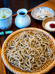 Cold soba noodles is presented in a woven bamboo basket. The meal is accompanied by dipping sauce, finely sliced green onion, wasabi, and side dish, creating a traditional Japanese dining experience