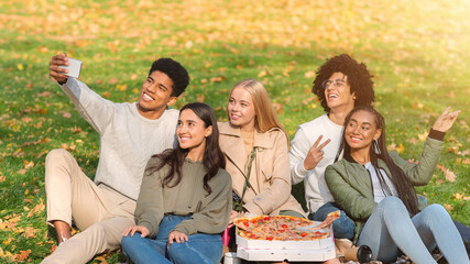 Cheerful multiracial teenagers having picnic in forest, taking selfie on smartphone, eating pizza