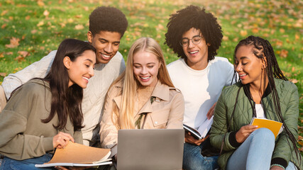Multiethnic cheerful friends making school homework while spending time in forest, looking at laptop screen