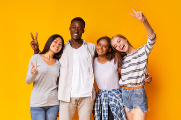 Carefree mixed race students having fun over yellow background, showing peace gesture