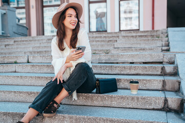 Young beautiful smiling woman in trendy summer clothes. Carefree female posing in street in sunny day. Positive model holds smartphone, looks at cellphone screen, uses mobile apps, in hat