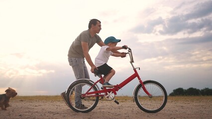 Obraz premium dad teaches son to ride a bike. happy family kid dream concept. the boy sat on bicycle for the first time, his father teaches boy to ride a bicycle. dog runs with family, fun family pastime lifestyle
