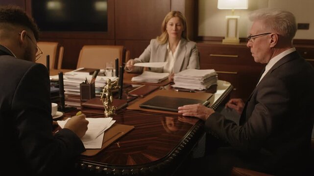 Zoom out of senior grey-haired attorney putting folder on wooden table in conference room, taking his place and opening folder, two colleagues preparing documents