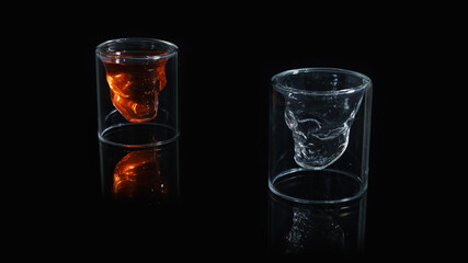 A pair of skull shaped shot glass, one filled with red orange drink and the other empty on a black background