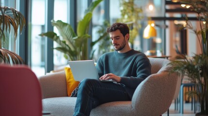Male office employee in a casual setting, working from a laptop in a lounge area, modern office with a relaxed atmosphere, blending work and comfort.