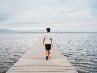 Rear view of a lonely young boy walking on a wooden pier surrounded by a calm body of water.