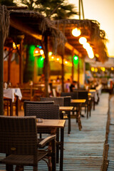 The terrace of a tavern on the beach, with wooden tables and chairs, after sunset. The atmosphere is animated by colored lights