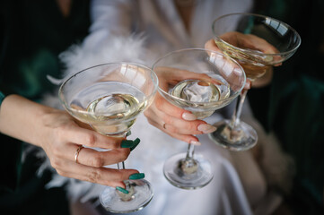 Bride and bridesmaid celebrate bachelorette spa party. Cheers. Pretty ladies friends wear silk robes hold glasses with champagne in hotel. Closeup three female hands holding cocktails, drink. Top view