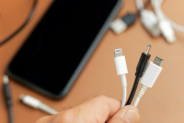 Hand holding a different type of electric charging cables, with phone and confused wire in the background .
