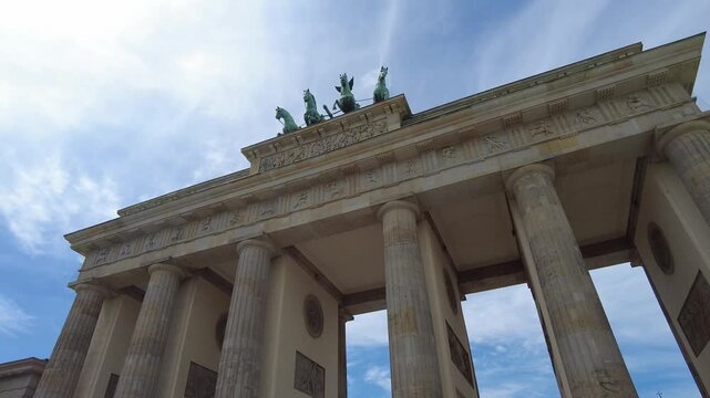 Perspective view from below the Brandenburg Gate. The statue of the horses atop the gate stand out against a blue sky with suggestive clouds. circled movement to the left of the camera
