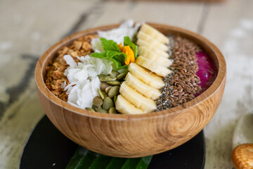 Healthy Dragonfruit Strawberry and Apple Smoothie Bowl topped with Banana Slices, Chia Seeds, Flax Seeds, Pumpkin Seeds, Coconut, Granola, Mint. In wooden bowl on rustic white paint table.