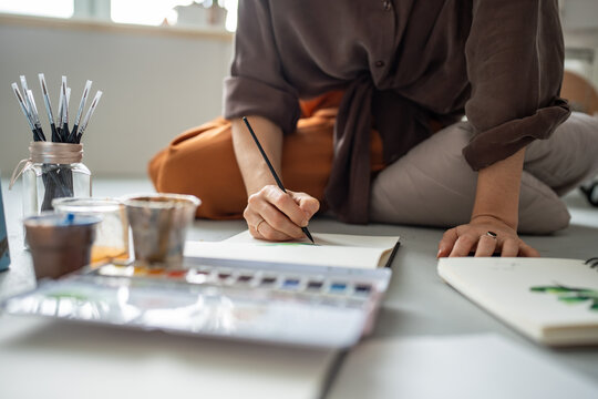 Closeup designer hands holding paintbrush painting with watercolors paints in sketchbooks, sits on floor in art studio. Creative occupation, making art orders. Student learning to draw in new technics