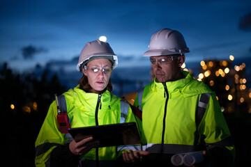Engineers wear uniform and helmet stand in holding tablet computer , survey inspection work plant site use radio communication to communication with night lights oil refinery background.