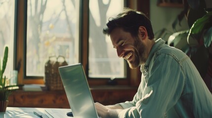 A man with a big smile leans forward over his laptop, bathed in natural light, working joyfully from a cozy, plant-filled home office.