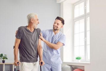 Fototapeta premium Happy health care worker with elderly man smiling and hugging in rehabilitation clinic. Young cheerful nurse helping senior patient to walk with his crutches. Physiotherapist helping old man in rehab