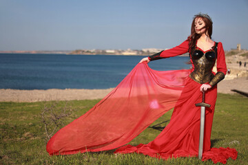 Fototapeta premium Beautiful red haired girl in metal medieval armor dress with sword standing in warlike pose