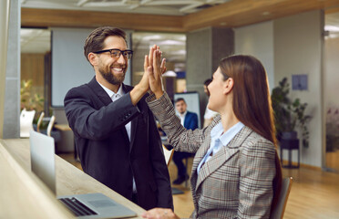 Two happy business people make a deal. Man and woman, sitting at an office table with a laptop computer, smile and give each other a high five. Cooperation, teamwork, success concept 