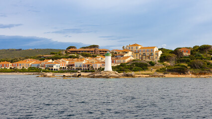 El faro de entrada al puerto de Palau, en Cerdeña, Italia, se alza sobre rocas que bordean la costa, rodeado de encantadoras casas de tonos pastel y techos de tejas rojas.