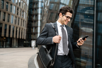 A businessman in a suit smiles as he checks his phone while walking through a modern urban setting,...