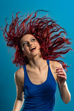 Portrait of a young happy dancing woman in long tight blue dress and long flying red wavy hair on a blue stdio background