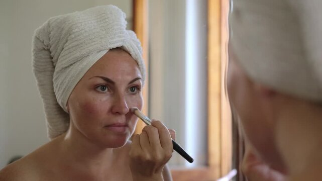 Woman does makeup in front of a mirror applies foundation and concealer with brush, after showering with a towel on her head
