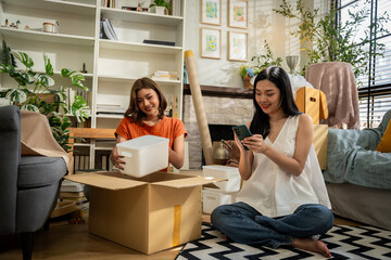 Two women sitting on a checkered rug in a room with boxes and books