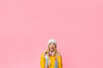 Amazed woman in white winter hat and yellow knitted sweater pointing up at copy space over yellow background