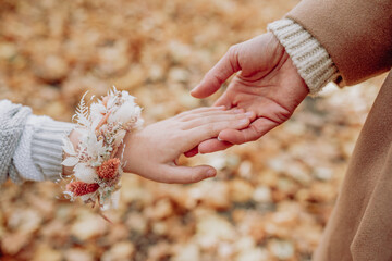 Close-up of two hands touching, one child wearing a floral bracelet and the other an adult, set against a backdrop of autumn leaves, capturing a tender, intergenerational moment filled