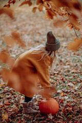 A child in a cozy autumn setting, wearing a warm orange sweater and grey beanie, bends down to pick up a pumpkin on a bed of fallen leaves..