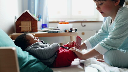 Older sibling gently holds the baby’s feet, both on a cozy bed in a sunlit room with toys and a gingerbread house on the table, showcasing a heartwarming moment of bonding and affection