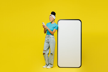 Full body side view young Latin woman wears blue top t-shirt headband casual clothes stand near big huge blank screen mobile cell phone with area using smartphone isolated on plain yellow background.